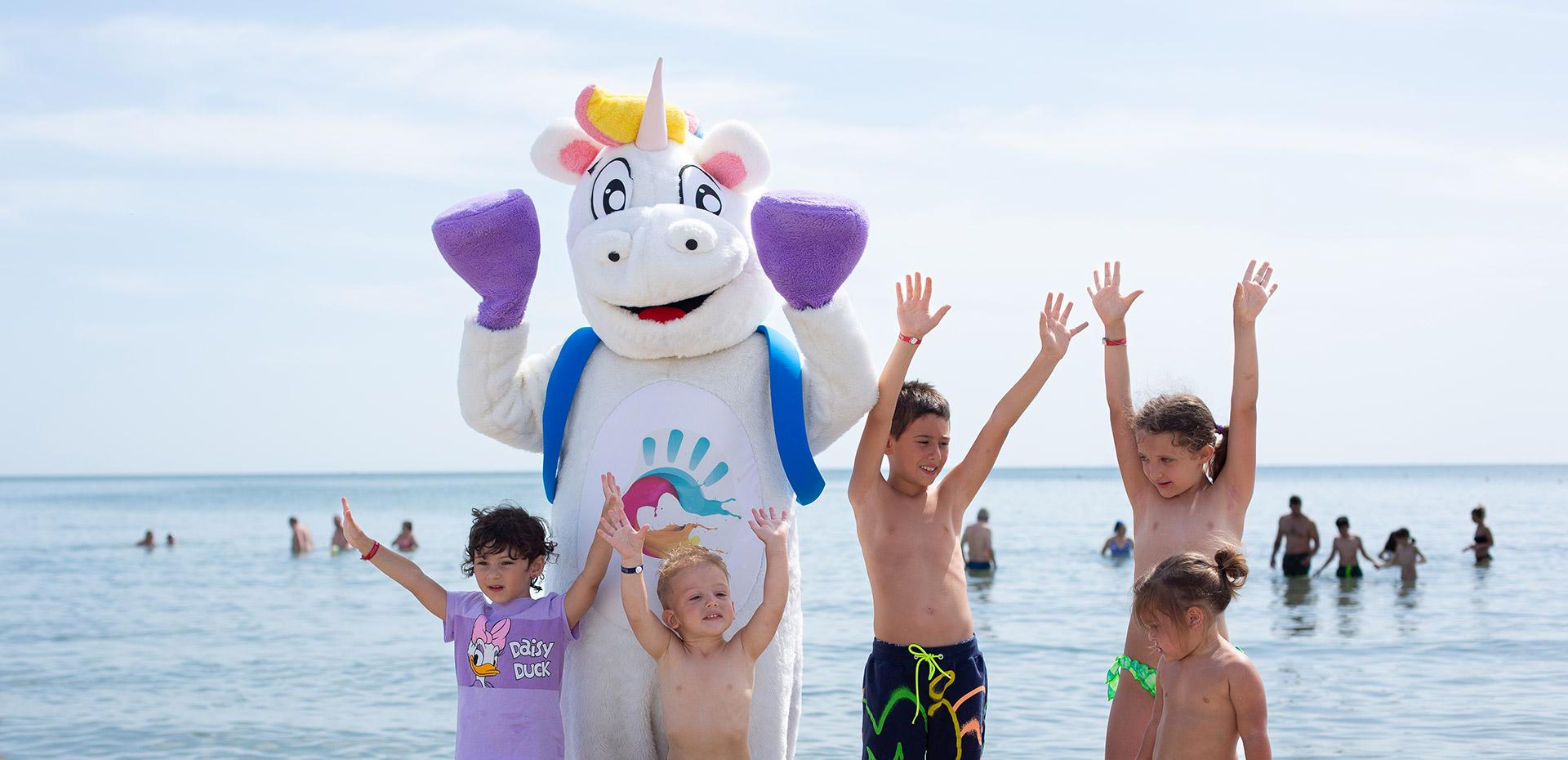 Fröhliche Kinder am Strand mit einem Einhorn-Maskottchen, Arme hoch.