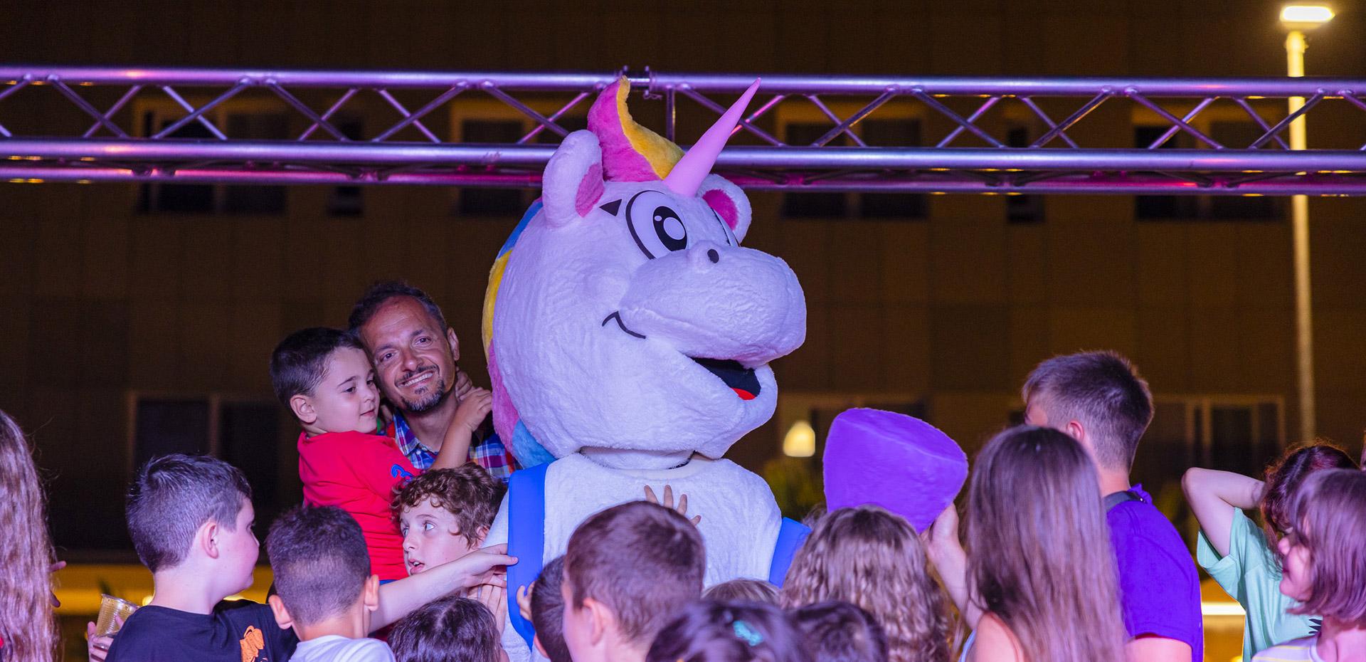 Fröhliche Kinder umarmen ein großes buntes Einhorn auf einer Party.