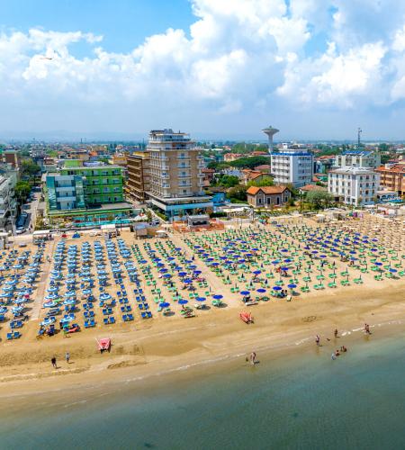 Plage bondée, parasols colorés, mer calme, bâtiments en arrière-plan, ciel bleu.