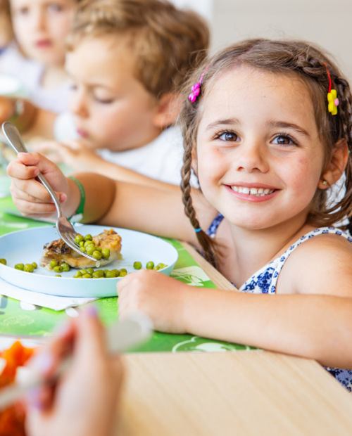 Bambina sorridente mangia un panino, con altri bambini sullo sfondo.