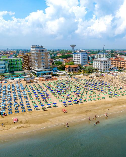 Spiaggia affollata, ombrelloni colorati, mare calmo, edifici sullo sfondo, cielo azzurro.